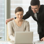 Female and male employees, office colleagues, business partners working together in office. Smiling businesswoman sitting at desk, typing on laptop, businessman standing near by and looking on screen